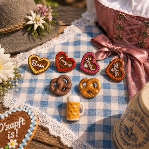 Könnte beinhalten: Eine Sammlung deutsch-thematischer Artikel auf einem blau-weiß karierten Tuch. Enthält herzförmige Lebkuchen mit Text, Brezeln, einen Miniatur-Bierkrug und einen traditionellen Hut mit einer Blume. Ein rosa Kleid mit einer Schleife ist im Hintergrund.