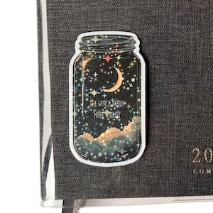 A whimsical sticker in the shape of a mason jar, filled with a night sky scene featuring a crescent moon, stars, and clouds. The text "THE WORLD NEEDS MORE WHIMSY" is visible. The sticker is placed on a dark gray textured journal.