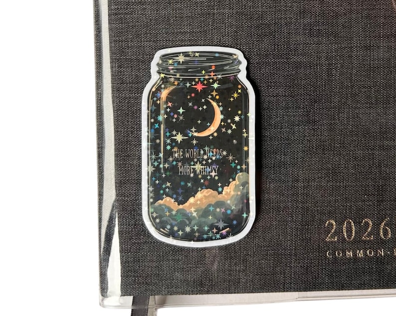A whimsical sticker in the shape of a mason jar, filled with a night sky scene featuring a crescent moon, stars, and clouds. The text "THE WORLD NEEDS MORE WHIMSY" is visible. The sticker is placed on a dark gray textured journal.