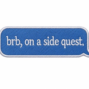 May include: A rectangular blue embroidered patch shaped like a speech bubble with rounded corners and a white border. The text "brb, on a side quest," is stitched in white lowercase letters across the center of the blue fabric surface.