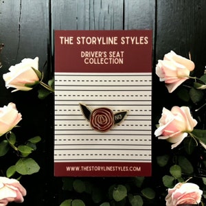 Puede incluir: Pin de esmalte dorado con una rosa roja con hojas negras y la palabra "NO" en el lado derecho. El pin est&aacute; en una tarjeta a rayas blancas y rojas con el texto "The Storyline Styles Driver's Seat Collection" y la direcci&oacute;n del sitio web "www.thestorylinestyles.com".