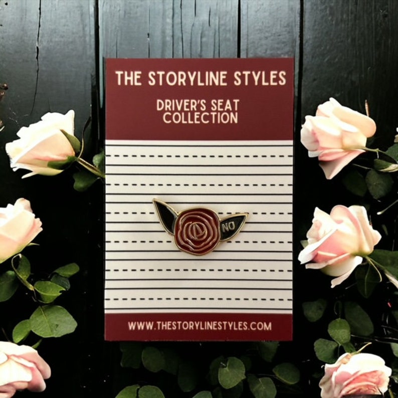 Puede incluir: Pin de esmalte dorado con una rosa roja con hojas negras y la palabra "NO" en el lado derecho. El pin est&aacute; en una tarjeta a rayas blancas y rojas con el texto "The Storyline Styles Driver's Seat Collection" y la direcci&oacute;n del sitio web "www.thestorylinestyles.com".