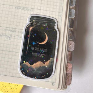 A sticker in the shape of a mason jar, filled with a night sky scene. The design includes a crescent moon, stars, and clouds, with the text "THE WORLD NEEDS MORE WHIMSY". The sticker is placed on a notebook page.
