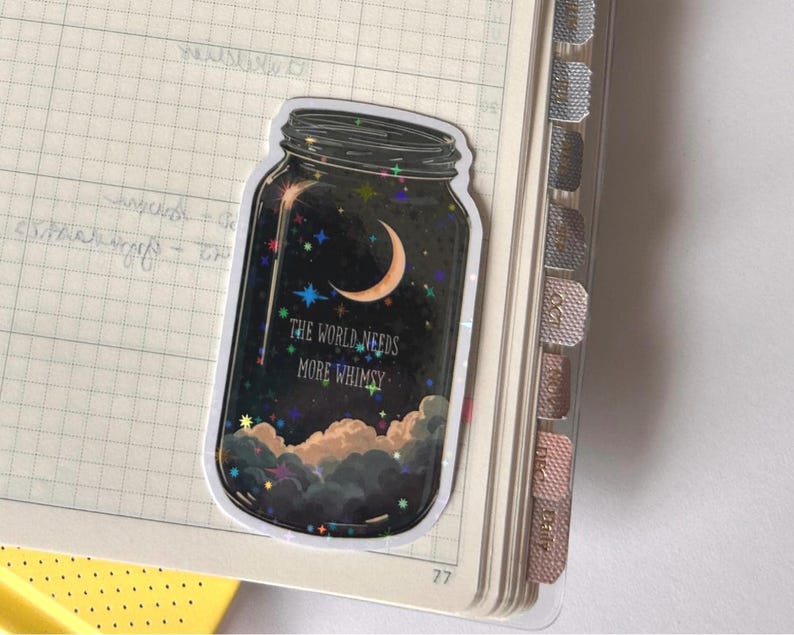 A sticker in the shape of a mason jar, filled with a night sky scene. The design includes a crescent moon, stars, and clouds, with the text "THE WORLD NEEDS MORE WHIMSY". The sticker is placed on a notebook page.