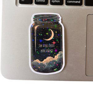 A jar-shaped sticker with a night sky design. The sticker features a crescent moon, stars, and clouds. Text on the sticker reads "THE WORLD NEEDS MORE WHIMSY". The sticker is placed on a laptop.