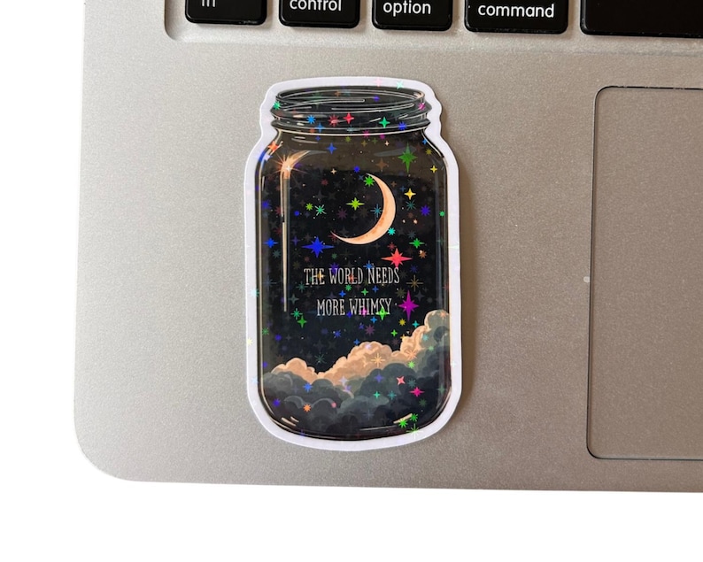 A jar-shaped sticker with a night sky design. The sticker features a crescent moon, stars, and clouds. Text on the sticker reads "THE WORLD NEEDS MORE WHIMSY". The sticker is placed on a laptop.