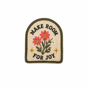 May include: An arched embroidered patch with a cream background and a dark green border. It features two pink flowers, small white blossoms, green leaves, and two yellow stars. The black text reads MAKE ROOM FOR JOY in a curved layout.