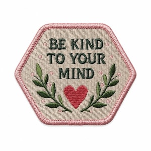 May include: A hexagonal embroidered patch with a beige background and a pink stitched border. The text BE KIND TO YOUR MIND is embroidered in dark green. Below the text is a red heart flanked by two green leafy branches with small pink dots.