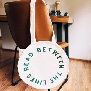 May include: White canvas tote bag with green text that reads "Read Between The Lines" in a circle around the bag.