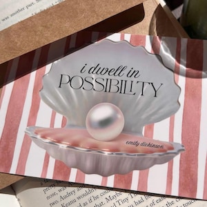 May include: A greeting card with a pearl inside a seashell illustration. The card has a pink and white striped background and the words "i dwell in POSSIBILITY" and "emily dickinson" printed on it. The card is resting on a book and a brown envelope.