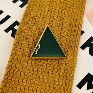 May include: A green enamel triangle pin with gold trim, reading "CHIN UP" in white lettering, is displayed on a textured mustard-yellow fabric. The pin is a geometric accessory.