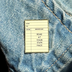 May include: A rectangular enamel pin resembling a library card, with the words "READ AT YOUR OWN PACE" in black text. The pin is set against a blue denim background.