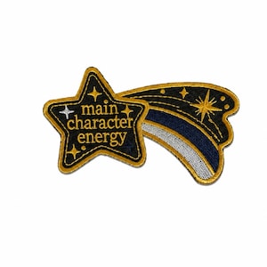 May include: A black and gold embroidered patch shaped like a shooting star. The star portion features the text "main character energy" in gold thread, surrounded by small gold and silver star icons. The tail is black, gold, and silver.