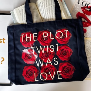 May include: A black canvas tote bag with white text that reads "The Plot Twist Was Love". The bag is decorated with red roses.