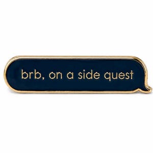 May include: A rectangular enamel pin with rounded corners shaped like a speech bubble. The pin features a dark navy blue background with gold-colored metal edges and the gold-colored text "brb, on a side quest" centered on the front surface.