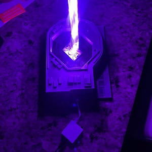 May include: A white 3D printed model of a futuristic device with a purple light emanating from the top. The device has a circular opening in the center and a series of buttons on the side. A white square-shaped object is connected to the device by a black wire.