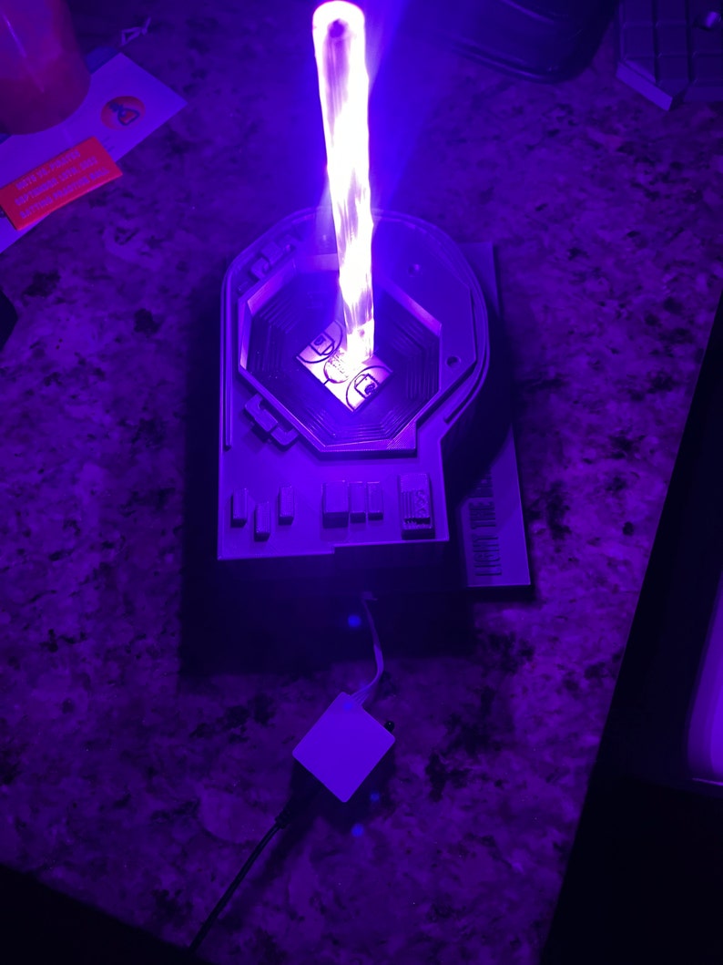 May include: A white 3D printed model of a futuristic device with a purple light emanating from the top. The device has a circular opening in the center and a series of buttons on the side. A white square-shaped object is connected to the device by a black wire.