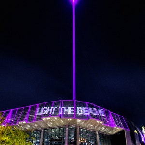 May include: A purple laser beam shoots up into the night sky above a large building with the words "LIGHT THE BEAM!" illuminated in purple on the side.