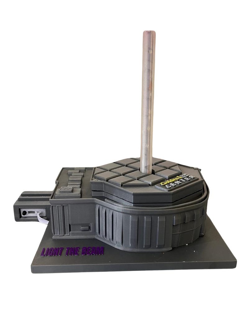 May include: A gray 3D printed model of a Star Wars lightsaber stand. The stand features a clear plastic lightsaber holder, a gray base with the text "Light the Beam" and a gray top with the text "Golden Tee Center".