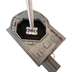 May include: A silver basketball hoop with a purple "LIGHT THE BEAM" inscription. The hoop is mounted on a silver pole and is surrounded by a black and white basketball court.