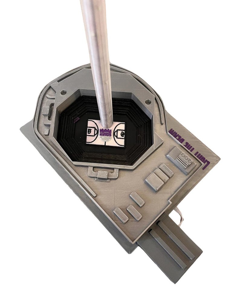 May include: A silver basketball hoop with a purple "LIGHT THE BEAM" inscription. The hoop is mounted on a silver pole and is surrounded by a black and white basketball court.
