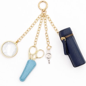 May include: A gold-toned keychain featuring four dangling charms: a circular magnifying glass, a pair of gold scissors in a light blue textured sheath, a small silver metal tool, and a cylindrical dark navy blue case with a gold zipper closure.