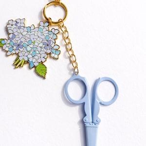 May include: A gold-toned keychain with a light blue pair of scissors and a floral charm. The charm features a bouquet of blue and purple flowers with green leaves. The scissors are a light blue color and are attached to the keychain with a gold chain.