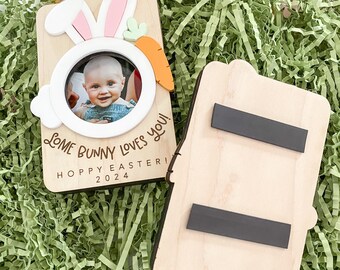 May include: A wooden photo frame shaped like a bunny with a carrot nose. The frame has a space for a photo and the text "Some bunny loves you! Hoppy Easter! 2024". The back of the frame has a magnetic strip.