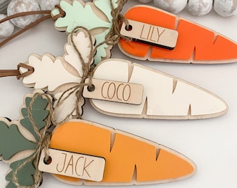 May include: Three wooden carrot ornaments with green tops and brown twine bows. Each carrot has a wooden tag with a name on it: Lily, Coco, and Jack.