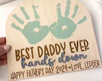 Puede incluir: Un letrero de madera con una huella de mano verde claro a cada lado. El letrero dice "Best Daddy Ever hands down Happy Father's Day 2024 - Love, Ledger".