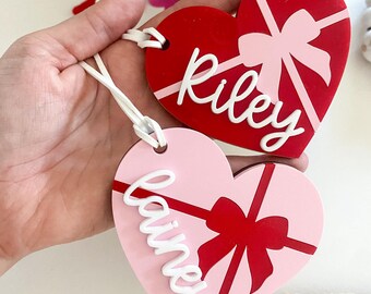 Könnte beinhalten: Zwei rote und rosafarbene herzförmige Geschenkanhänger mit weißem Band. Die Anhänger haben eine weiße Schleife und die Namen "Riley" und "Rainier" in weiß.