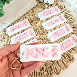 May include: Personalised keyrings with names in pink, featuring ballet shoes. The keyrings are rectangular with a white background and gold heart-shaped clasps. They are displayed on a woven mat.