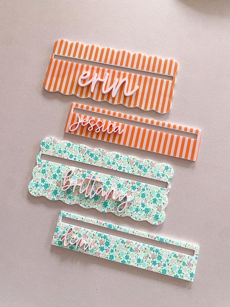 K&ouml;nnte beinhalten: Vier personalisierte Namensschilder. Zwei sind orange und wei&szlig; gestreift mit den Namen "erin" und "jessica". Die anderen beiden sind floral mit den Namen "brittany" und "ken". Jede Karte hat einen gewellten Rand.
