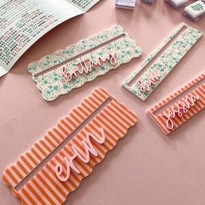 K&ouml;nnte beinhalten: Vier personalisierte Namensschilder. Zwei sind floral gemustert mit rosa Namen, eines ist orange gestreift mit einem rosa Namen und eines ist orange gestreift mit einem rosa Namen. Mahjong-Steine und ein Papier mit Text befinden sich im Hintergrund.