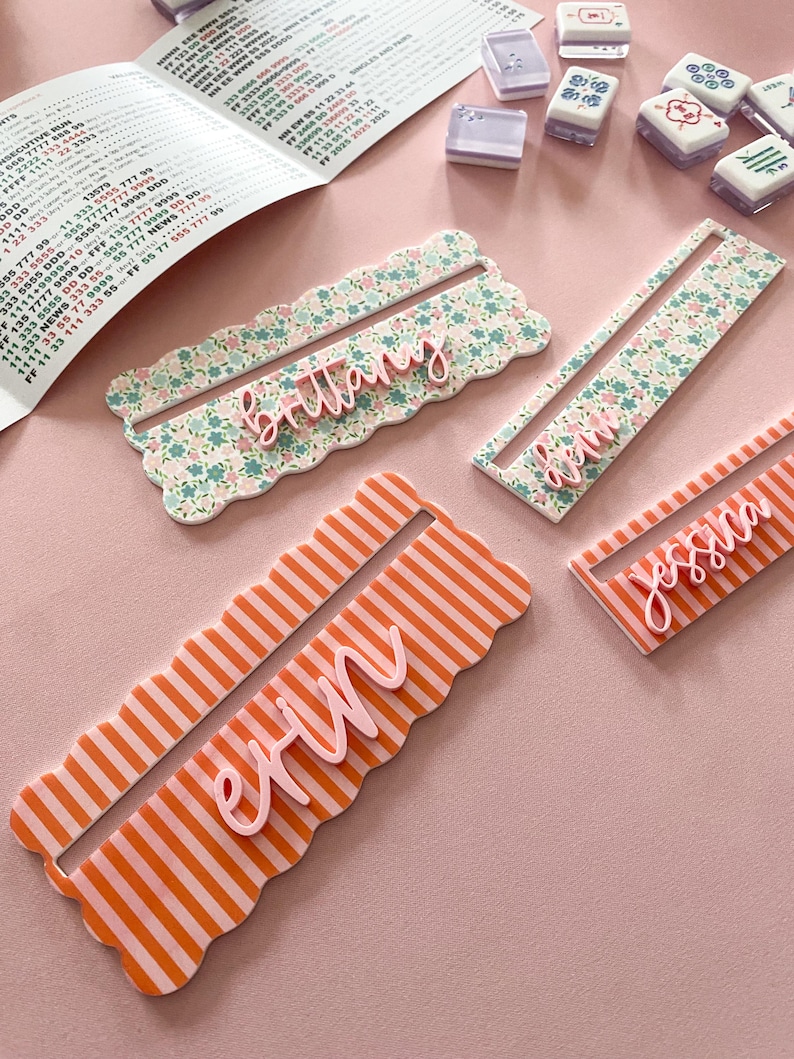 K&ouml;nnte beinhalten: Vier personalisierte Namensschilder. Zwei sind floral gemustert mit rosa Namen, eines ist orange gestreift mit einem rosa Namen und eines ist orange gestreift mit einem rosa Namen. Mahjong-Steine und ein Papier mit Text befinden sich im Hintergrund.