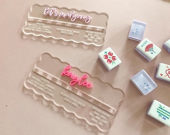 May include: Clear acrylic Mahjong name card holders with pink lettering that reads "let's mahjong" and "Kaylee". Several Mahjong tiles are scattered around the holders, featuring various designs and colors.