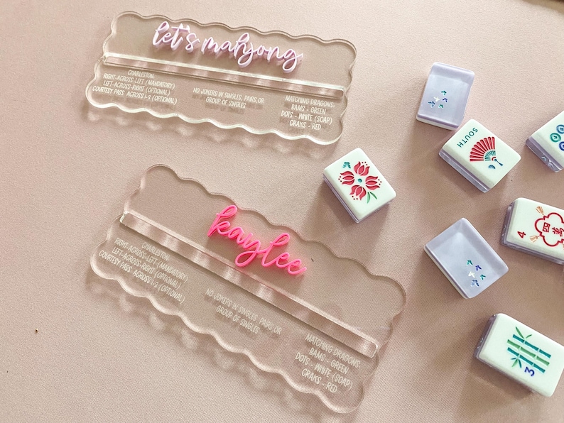 May include: Clear acrylic Mahjong name card holders with pink lettering that reads "let's mahjong" and "Kaylee". Several Mahjong tiles are scattered around the holders, featuring various designs and colors.