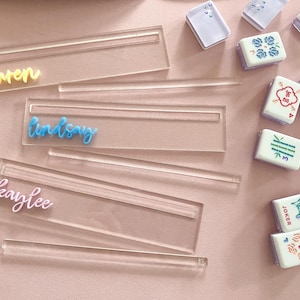 May include: Clear acrylic place card holders with names in yellow, blue, and pink script. Mahjong tiles with various designs are scattered around the place cards on a pink surface. The place cards have a slot for the name cards.