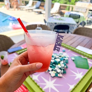 K&ouml;nnte beinhalten: Ein rosa Cocktail in einem transparenten Plastikbecher mit einem roten Strohhalm. Der Becher hat ein Namensschild mit der Aufschrift "KAYLEE". Das Getr&auml;nk wird &uuml;ber einem Tisch mit einem lila-wei&szlig;en Sternmuster gehalten.