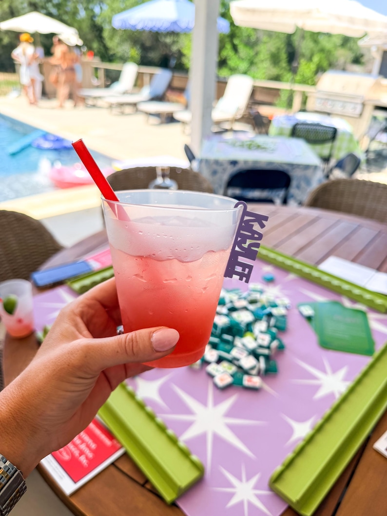 K&ouml;nnte beinhalten: Ein rosa Cocktail in einem transparenten Plastikbecher mit einem roten Strohhalm. Der Becher hat ein Namensschild mit der Aufschrift "KAYLEE". Das Getr&auml;nk wird &uuml;ber einem Tisch mit einem lila-wei&szlig;en Sternmuster gehalten.