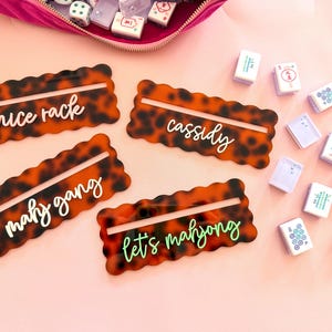Op de afbeelding: Vier Mahjong-tegelhouders met schildpadpatroon met witte en groene tekst. De tekst luidt "nice rack", "cassidy", "mahj gang" en "let's mahjong". Mahjong-tegels en een roze fluwelen etui staan ook op de afbeelding.