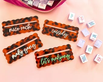 May include: Four tortoise-shell-patterned Mahjong tile holders with white and green text. The text reads "nice rack", "cassidy", "mahj gang", and "let's mahjong". Mahjong tiles and a pink velvet pouch are also in the image.