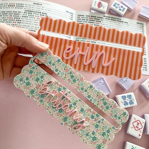 K&ouml;nnte beinhalten: Zwei personalisierte Mahjong-Kartenhalter. Einer ist orange mit wei&szlig;en Streifen und dem Namen "Erin" in Rosa. Der andere ist floral mit dem Namen "Brittany" in Rosa. Mahjong-Steine sind um die Halter herum verstreut.