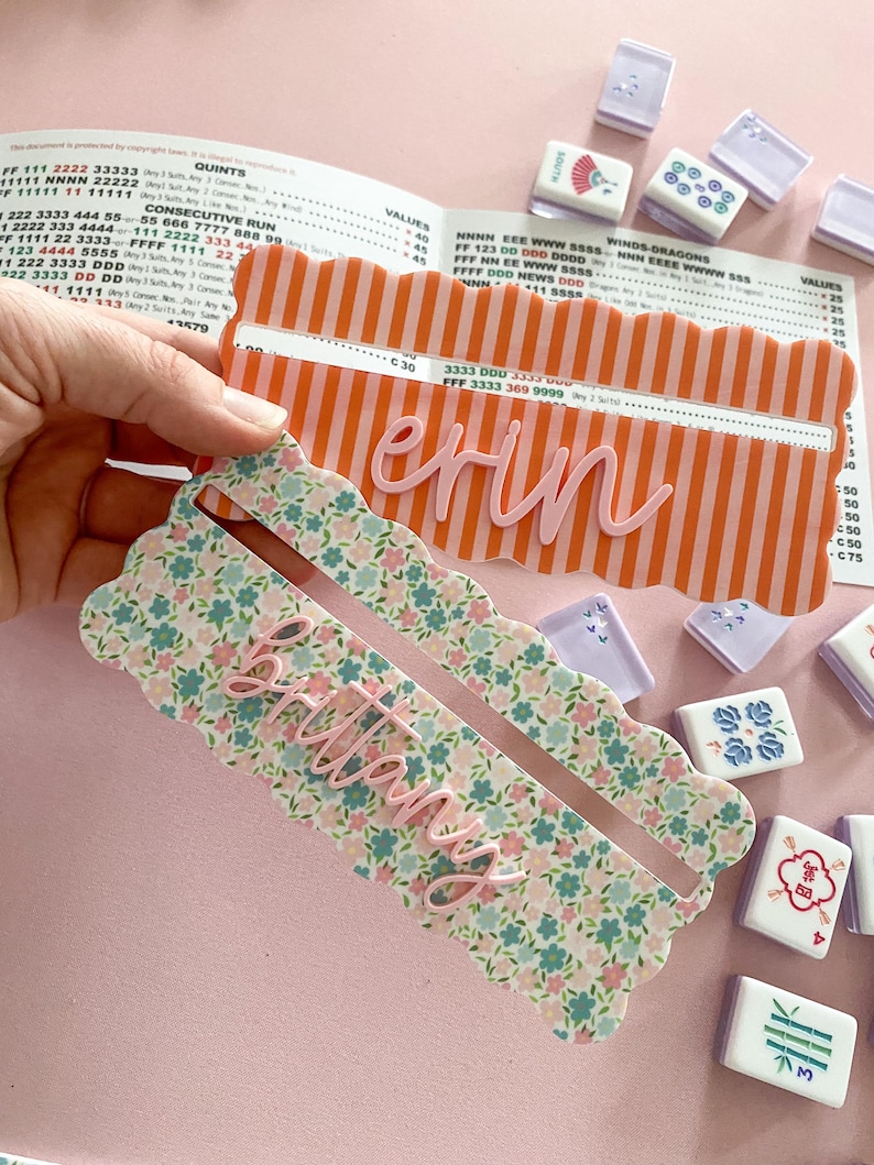 K&ouml;nnte beinhalten: Zwei personalisierte Mahjong-Kartenhalter. Einer ist orange mit wei&szlig;en Streifen und dem Namen "Erin" in Rosa. Der andere ist floral mit dem Namen "Brittany" in Rosa. Mahjong-Steine sind um die Halter herum verstreut.