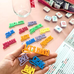 K&ouml;nnte beinhalten: Eine Ansammlung farbenfroher Namensschilder und Spielsteine auf einer rosa Oberfl&auml;che. Die Schilder, in verschiedenen Farben, schreiben Namen und Phrasen. Ein klares Glas und eine rosa Tasche sind ebenfalls vorhanden.