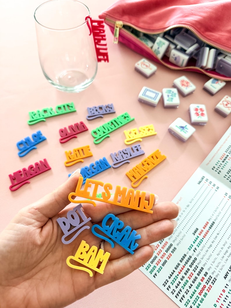 K&ouml;nnte beinhalten: Eine Ansammlung farbenfroher Namensschilder und Spielsteine auf einer rosa Oberfl&auml;che. Die Schilder, in verschiedenen Farben, schreiben Namen und Phrasen. Ein klares Glas und eine rosa Tasche sind ebenfalls vorhanden.