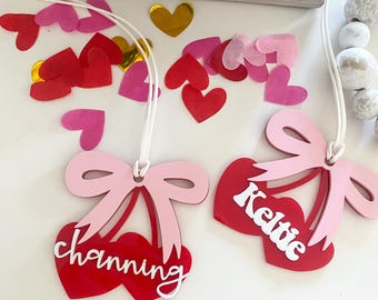Könnte beinhalten: Zwei rote kirschförmige Ornamente mit rosa Schleifen und weißen Buchstaben. Die Ornamente hängen an einer weißen Schnur und sagen "Keltie" und "channing".