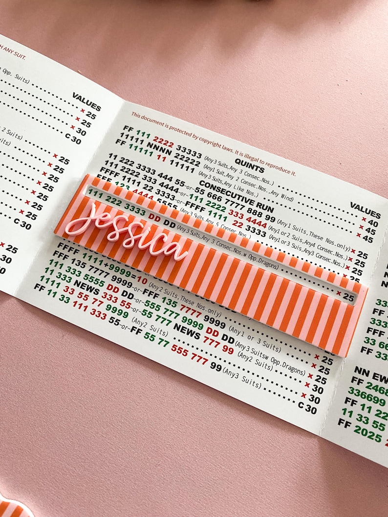 K&ouml;nnte beinhalten: Ein personalisiertes Namensschild aus Acryl mit dem Namen "Jessica" in wei&szlig;er Schrift, vor einem Hintergrund aus orangefarbenen und wei&szlig;en Streifen. Das rechteckige Schild liegt auf einem bedruckten Dokument.
