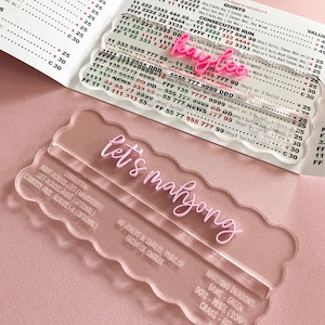 May include: Clear acrylic Let's Mahjong game board with pink text, alongside a pink name tag that says "Kaylee". The board includes instructions for play and a list of matching dragons, bams, dots, and craks.