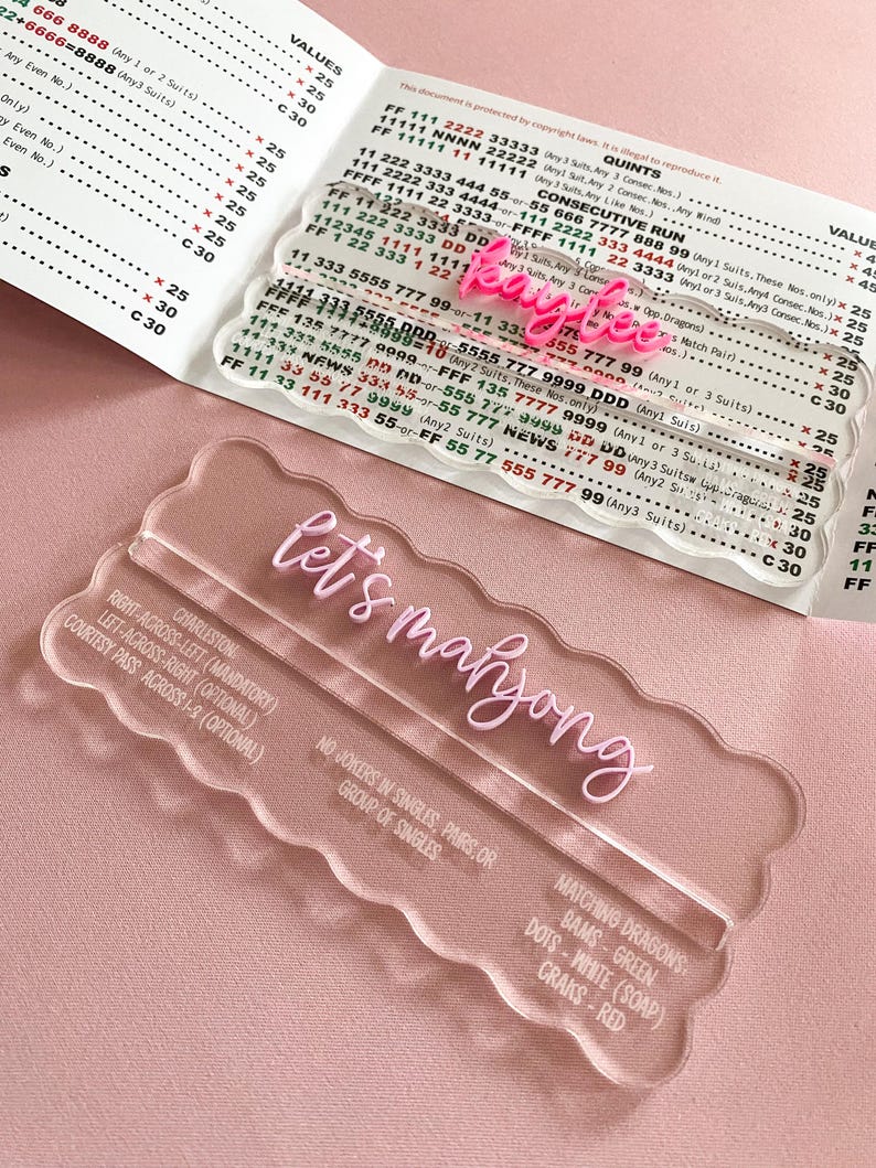 May include: Clear acrylic Let's Mahjong game board with pink text, alongside a pink name tag that says "Kaylee". The board includes instructions for play and a list of matching dragons, bams, dots, and craks.